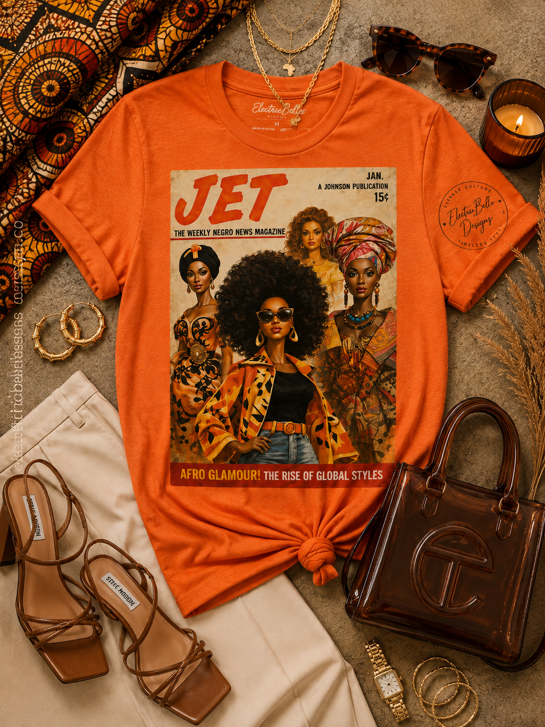 Flat lay of an orange JET Afro Glamour graphic t-shirt styled with cream trousers, brown strappy heels, a brown jelly bag, gold jewelry, tortoise sunglasses and African print accents.